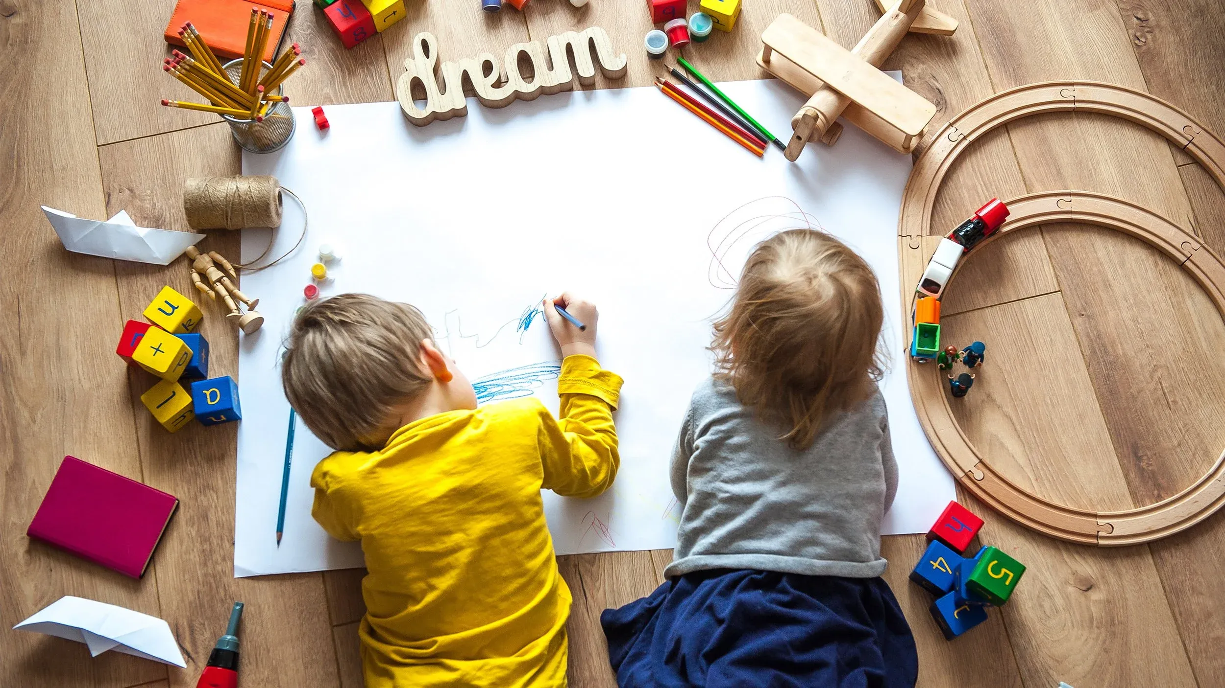 Fëmijë duke vizatuar në dysheme, të rrethuar nga lodra.